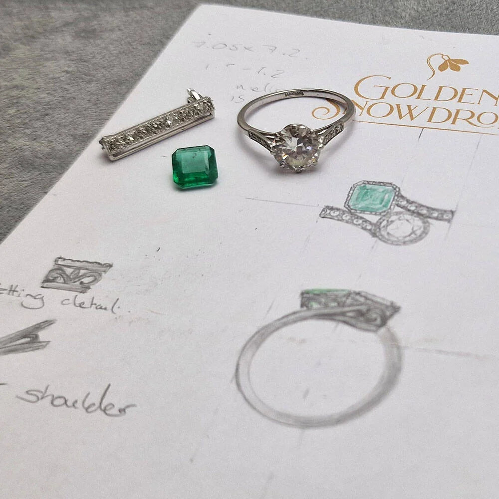 Flat-lay of jewellery design elements on a sheet of paper with hand-drawn ring sketches and the Golden Snowdrop logo. In the centre is a loose square-cut Zambian emerald gemstone. To the right is a classic diamond solitaire ring with a round brilliant centre stone. To the left is a straight bar of small diamonds set in metal. The sketches show a ring design incorporating the emerald